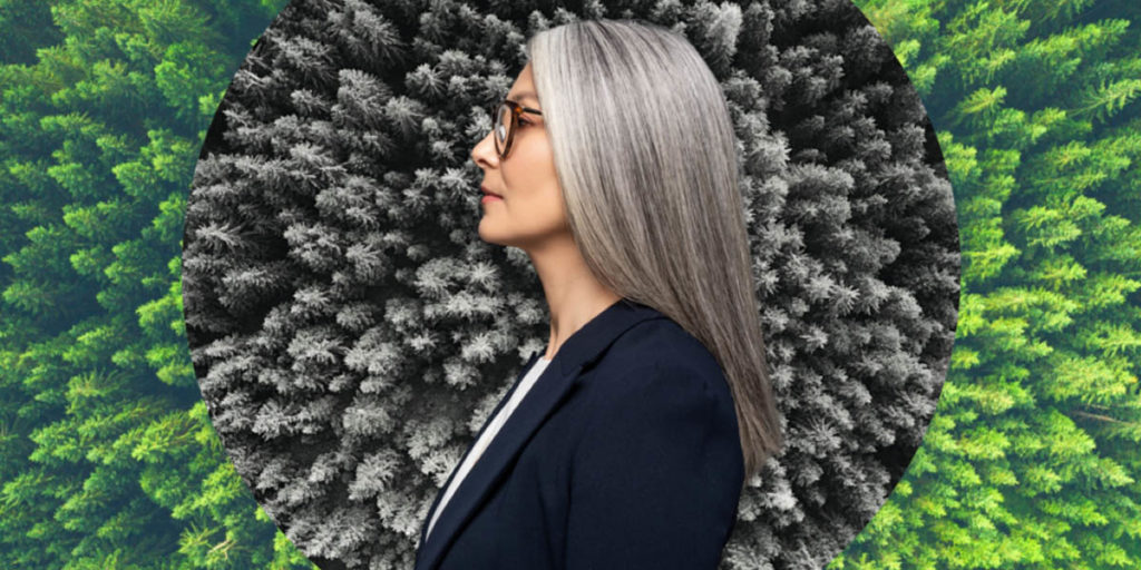 a grey haired white woman stands in front of green and grey trees